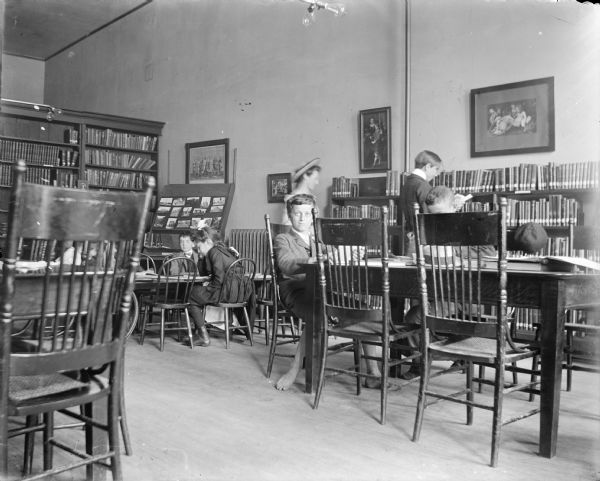 Children and young adults read in the public library. The boy facing the camera is barefoot. Bookshelves line the walls; prints and photographs are also on display.