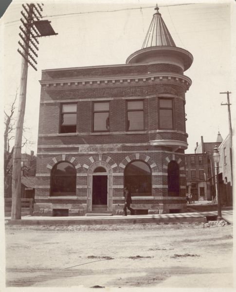 A man walks by the Madison police station, a two-story brick building with corner turret on the corner of South Webster Street. The fire station was across the alley to the right.