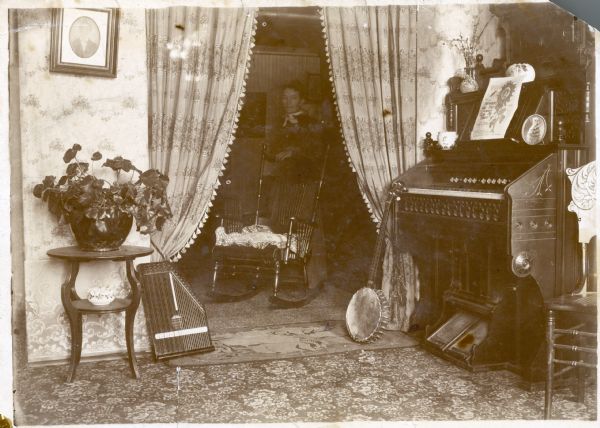 Clara Case Middleton stands behind a rocking chair beyond a curtained doorway. In the front room, a zither and banjo are displayed near a parlor organ.
