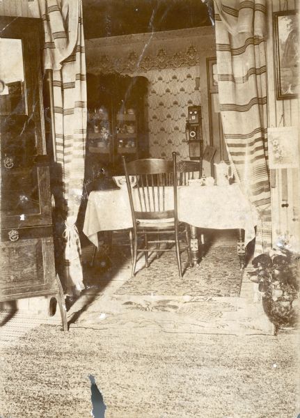A view through a curtained doorway into the Middleton family dining room at 22 East Mifflin Street. A decorative bowl and china coffee set are on the table. There is a telephone on the wall. A hooked rug featuring a lion and dated 1896, lies in the doorway.