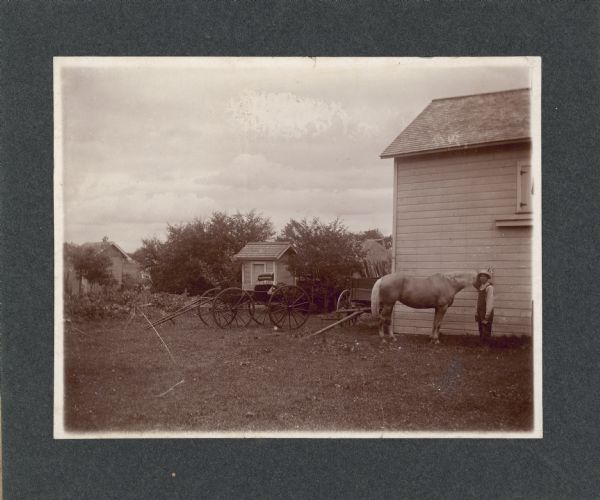 A man stands with a horse near a small barn. A buggy and wagon are parked nearby. There are two small sheds or outhouses near a garden plot and a haystack in the background.