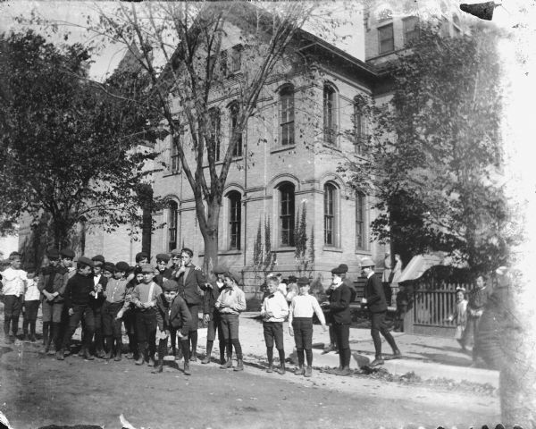 A group of boys, most wearing caps, pose at the side of Madison's Third Ward School, also known as Brayton School. There are girls in the background.