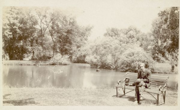 Forest Middleton sits with his camera beside him on a park bench near the lagoon in Tenney Park. An arched bridge is visible through the trees.