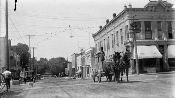 A man standing in a wagon drives a team of horses through an intersection. The horses are wearing horse fly-nets. A second man and dog walk on the left. There are automobiles parked along the street.