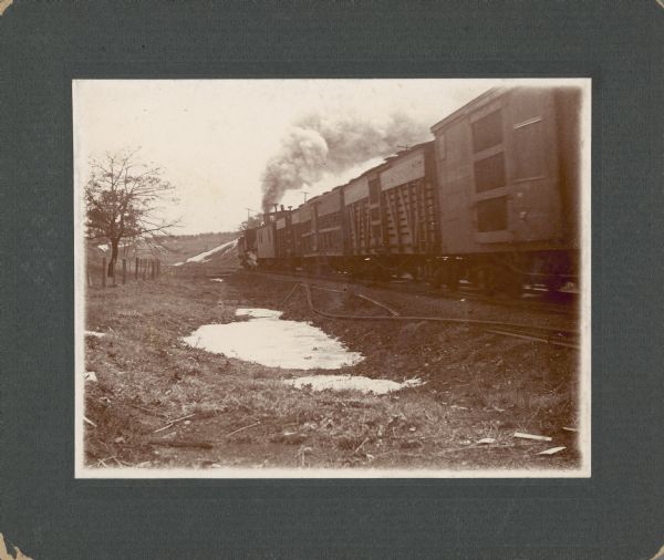 Smoke rises from a steam engine pushing (or pulling) a freight train. There are bent rails and snow in the ditch.