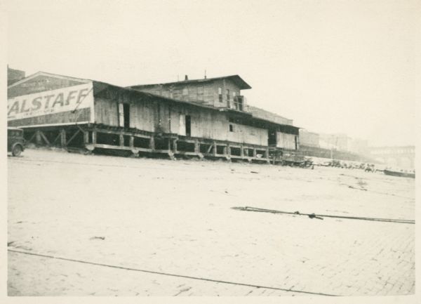 A weathered building stands on a beach, possibly in the state of Washington. A Falstaff beer sign is painted on its side. There is a bridge or viaduct in the far background. Several cars are parked on the beach.