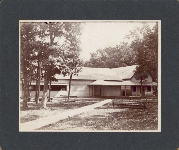 Two frame structures connected by an open porch stand in a wooded area. The building on the right appears to be a small two-story house. On the left is a one story structure with wooden awnings covering a row of screened windows. There are long benches on the lawn. A board sidewalk leads to the porch and double doors.
