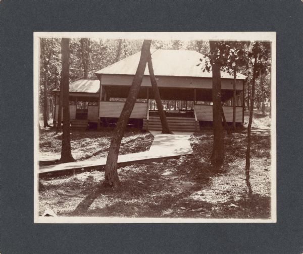 Benches are visible inside a wooden, open air pavilion with a hip roof and large doorway. There is a small attached porch on the left which mimics the shape of the main building. There is a large board sidewalk leading through the trees to the steps.