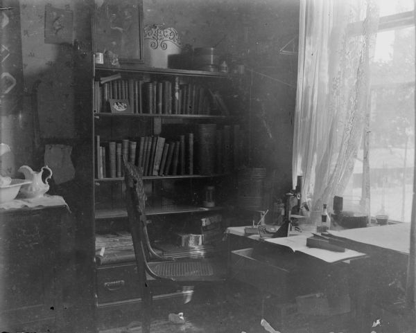 A small microscope sits on a table near a lace-curtained window in Forest Middleton's room. There is also a chair, bookcase and commode with china commode set in the room.