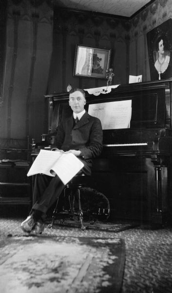 Forest Middleton, wearing eyeglasses and holding a musical score, sits with his back to an upright piano.