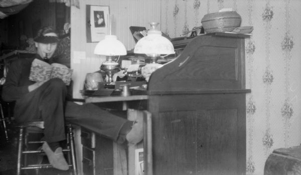 Forest Middleton, wearing a visor and moccassins, sits at a roll top desk. He holds an open book and is smoking a pipe. There are two oil lamps on the desk.