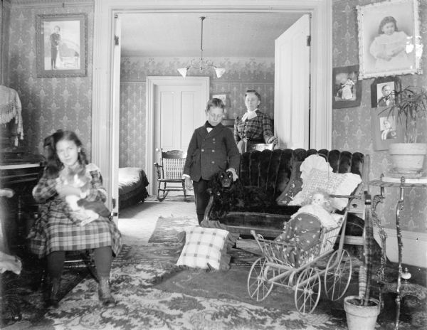 A girl on a rocking chair holds a rabbit, and a boy and a woman stand in the background. The boy rests his hand on a dogs head; the dog is lying on a love seat. There is a doll in a toy baby carriage on the right.