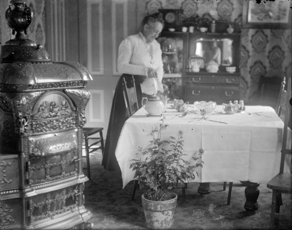 A woman poses as if pouring water into a glass. There is a tablecloth, tea set and glass bowl on the table. A large, fancy parlor stove is featured in the left foreground. A potted chrysanthemum has been placed near the table. In the background is a china cabinet; an indistinct image of the photographer is visible in the china cabinet's mirror.