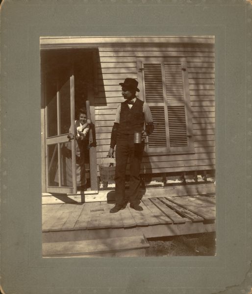 In a posed scene, the photographer, wearing sleeve protectors, stands on the porch holding a bucket and large measuring cup while his smiling wife Clara threatens him with a rolling pin. She is standing in the doorway, holding the screen door open.  Shutters cover a window.