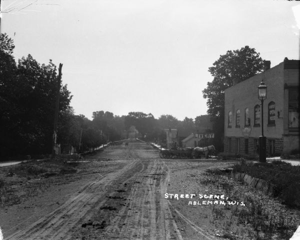 View features a dirt street and a horse-drawn wagon. On the right is a brick building with signs, a lamppost, and a sign that says "LIVERY". In the far background the street leads to what appears to be a bridge with an arch.