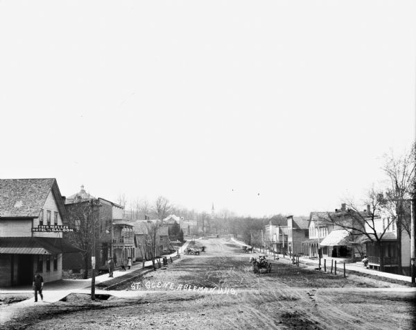View features a dirt street, horse-drawn wagons, storefronts, and several pedestrians walking on the sidewalk bordering the street. In the background is a hill with what appears to be a church steeple.