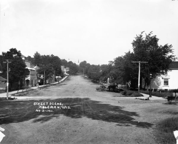 View features a dirt street, dwellings, a horse-drawn vehicle, sidewalks, power lines, and many trees. In the background is a hill with what appears to be a church steeple.