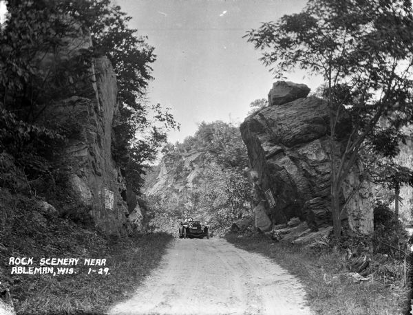 View of a man driving an automobile on a dirt road in Ableman. Rock formations are on both sides of the road, and rocks and trees are in the background.