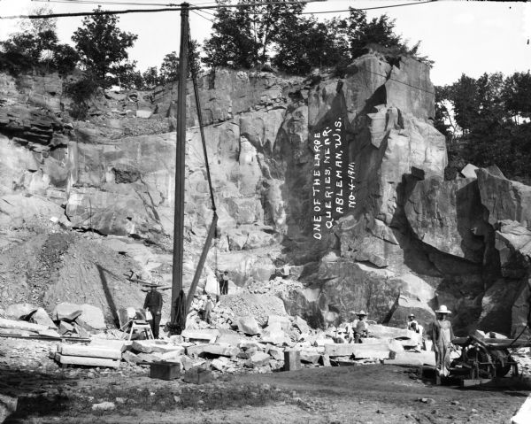 View of workers in a large quarry in Ableman. Several male workers are standing in the quarry among piles of rock with their work equipment.