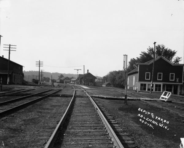 View down railroad tracks of a depot and yards in Ableman. The depot is in the background, among other industrial and commercial buildings.