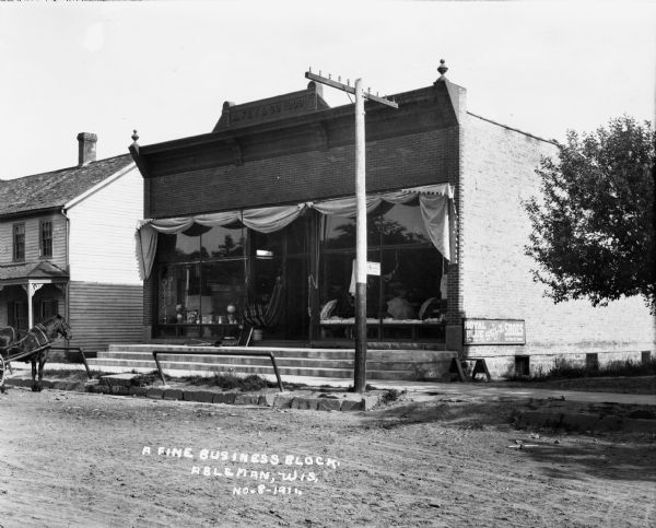 View from street of a business block in Ableman. There is a storefront with umbrellas displayed in the show window, and what appears to be a hammock is hanging near the front entrance. On the side of the building is a sign that says: "Royal Blue Shoes". A horse is hitched near the curb, pulling a wagon or carriage.