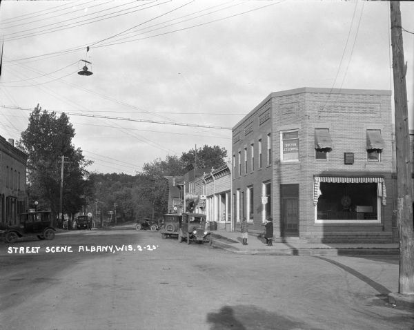 View down street. On the right on the corner is a bank, with the office of "Doctor J.T. Lemmel" on the second floor. Three children are standing on the sidewalk outside the bank entrance. Automobiles are parked on both sides of the street, and power lines and a streetlight are suspended over the street.