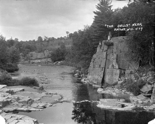 Elevated view of The Dells. A group of people are standing on a rock formation on the right overlooking the river. On the left is a rocky shoreline, and in the distance are trees and rock faces.