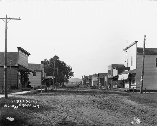 View down street of central business district. Storefronts and building facades are on both sides of the street, and a few pedestrians are standing or walking near the entrances to some of the buildings. There is a hill in the far background.