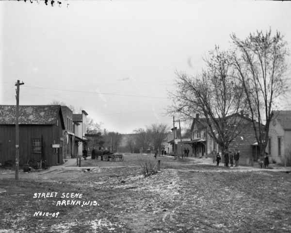 View down a dirt road bordered by several buildings. Pedestrians are standing on wooden sidewalks on both sides of the street. There is also a horse-drawn vehicle, and many trees and power lines. A hill is in the far background.
