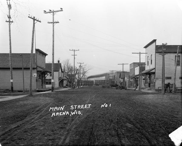 View down of Main Street, which is a dirt street passing through the central business district of the town, featuring several storefronts with porches and signs. An automobile and several horse-drawn vehicles are parked near the sidewalk. In the background a train is passing across the road, and in the far background is a hill.