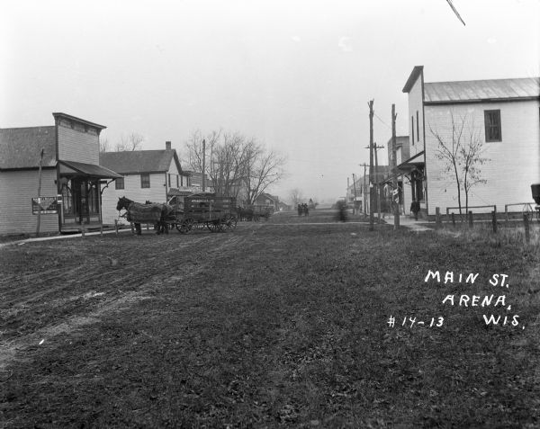 View down Main Street, which is a dirt street passing through the central business district of the town. There are several storefronts with signs and porches along a sidewalk. Several horse-drawn vehicles are hitched nearby, and pedestrians and other horse-drawn vehicles are in the background.