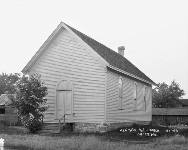 Exterior view of the front and side of the German Methodist Evangelical Church, which is a simple building with arched windows, a chimney, and wooden steps leading up to the entrance. Other buildings are in the background.