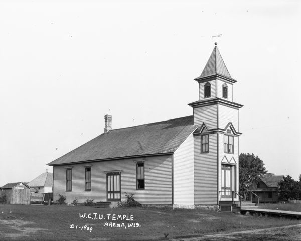 Exterior view of the Women's Christian Temperance Union (W.C.T.U) Temple. The temple is a simple building with a tower with a weather vane above the main entrance, and an elevated wooden walkway leading to the front steps. There is also a side entrance. A number of other buildings are in the background.