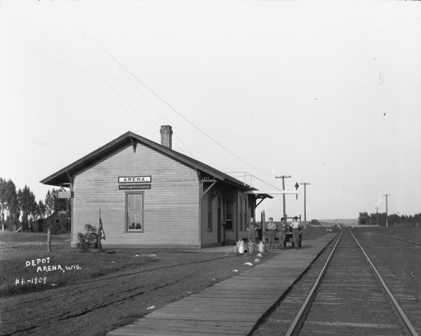 Exterior view from railroad tracks of the Depot. Four men are standing alongside the depot building near a wheeled cart. The building features two signs, one that says "ARENA," and the other that says "Wells Fargo & Co. Express." There is also a hand water pump on a landing near the depot on the left.