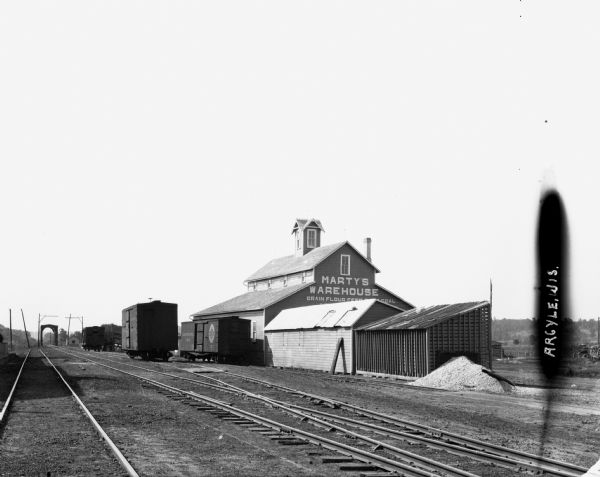 View of Marty's Warehouse from across railroad tracks. The warehouse building has large signs of painted lettering advertising the storage facilities. Railroad cars are sitting on the tracks to the left of the building.