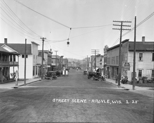 View of a street scene featuring a central business district bordered by sidewalks and storefronts. A number of people are standing on the sidewalk and walking in the background. Automobiles and horse-drawn wagons are parked on the side of the street. On the left is a barber pole in front of a building with a front porch and a balcony.