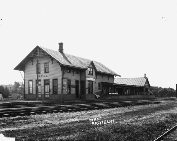 View across railroad tracks of the depot, which is a two-story building with a covered platform. There appears to be a commercial building next door on the right, and in the far background on the left is a barn.