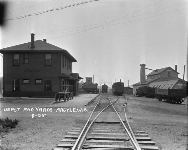 View down railroad tracks of a locomotive pulling into a railroad depot and yards. The depot is a two-story building. Two wagons are parked near the depot platform. There is a warehouse off to the right, and several railroad cars are sitting on a side track.