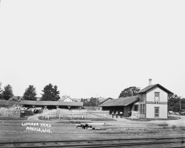 Elevated view across railroad tracks of a lumber yard. A two-story building on the right has a sign that says: "Lovejoy & Ringham". There are fences, a storage area for lumber, and a number of men walking in the yard. In the far background are houses and barns on a hill.