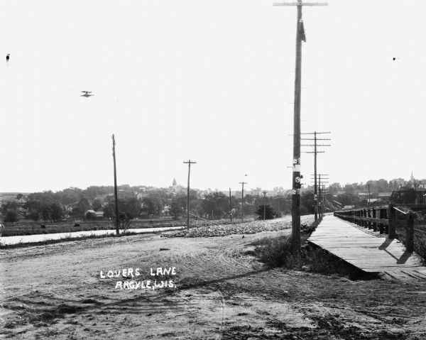 View of Lovers Lane, which a dirt road with a sidewalk and bridge on the right. There are power lines in the foreground, and a town in the background. There is a field off to the left, across what appears to be a river, with grazing animals.