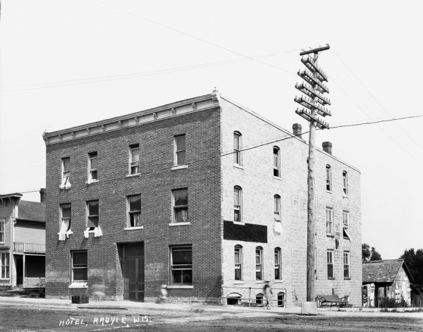 View from across street of a hotel with a brick facade. Many of the windows are open and the curtains are hanging out. There is a pedestrian walking on the sidewalk in front of the building near large power lines and a bench. In the background on the right is a fence and an old shed or barn plastered with posters and signs.