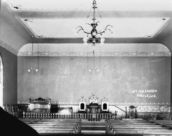 Interior view towards the front of the Methodist Evangelical Church. There is an altar flanked by two chairs, with an organ on the left. In the foreground are two rows of pews, and a large chandelier hangs from the ceiling.