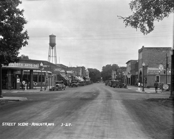 View down street. Cars are parked on either side. A repairman checks the tires of a car in front of the Augusta Auto Company, gas station and auto repair. Other stores and shops pictured are: the Grand Cafe, a pharmacy, Cameron's Candy, Horton Cafe, and the Augusta Barber Shop. A water tower is in the background.