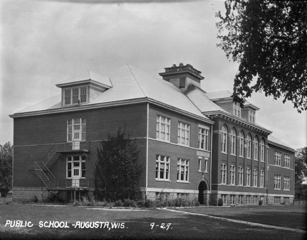 Exterior view of public school. There is a two-story fire escape on the side of the building. The words "High School" are over the arched entry at the front of the building.
