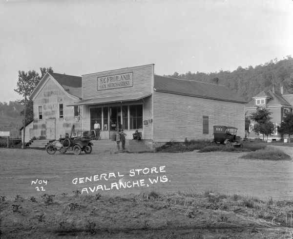 Exterior view of S.C. Froiland General Merchandise. A car with a young boy and girl is parked in front of the store, and another car is parked on the side of the building. Three men pose on the front steps of the store.
