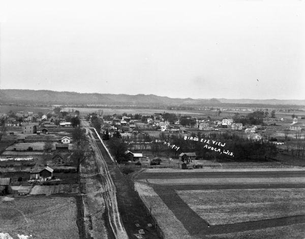 An elevated view of a town in the Wisconsin River Valley. Fields surround the town, and low hills are in the far background.
