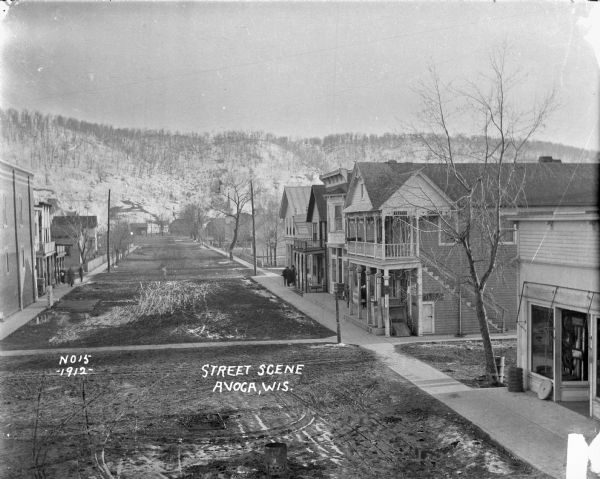 Elevated view of a wide main street through town. Pedestrians are on sidewalks on both sides of the dirt street. A church steeple is visible in the background near dwellings, and behind it are snow-covered steep hills.