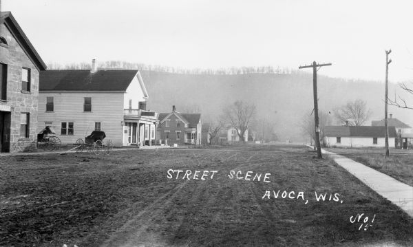 View down street of with commercial buildings and dwellings. There are two carriages parked near a building which has part of a sign visible over the entrance that says: "barn." There is a hill in the background.
