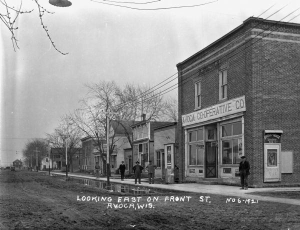 View of Front Street with five men standing along a sidewalk. They are standing near a two-story building which houses the Avoca Cooperative Company and the opera house. Caption reads: "Looking East on Front St., Avoca, Wis."