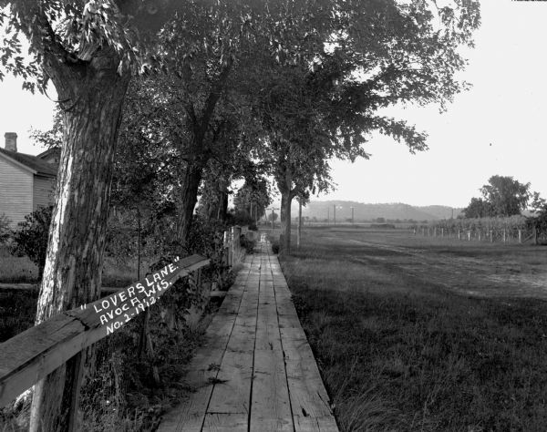 View down wooden walkway with a dwelling, trees and backyards on the left and an open area on the right.